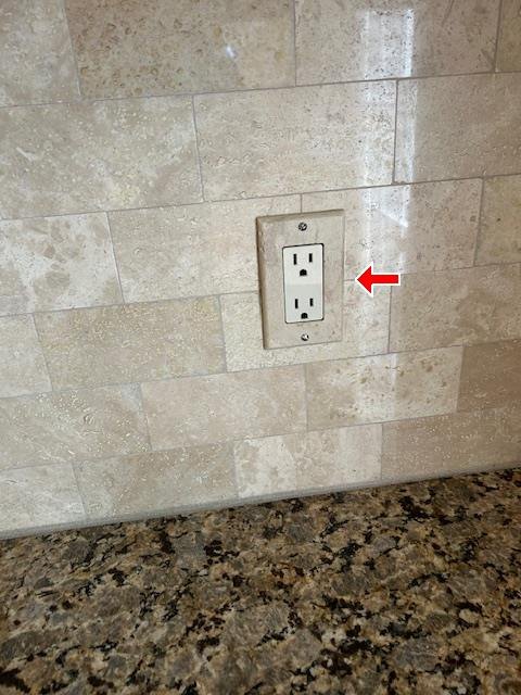 Electrical outlet in a stone tile backsplash above a granite countertop, red arrow pointing at it