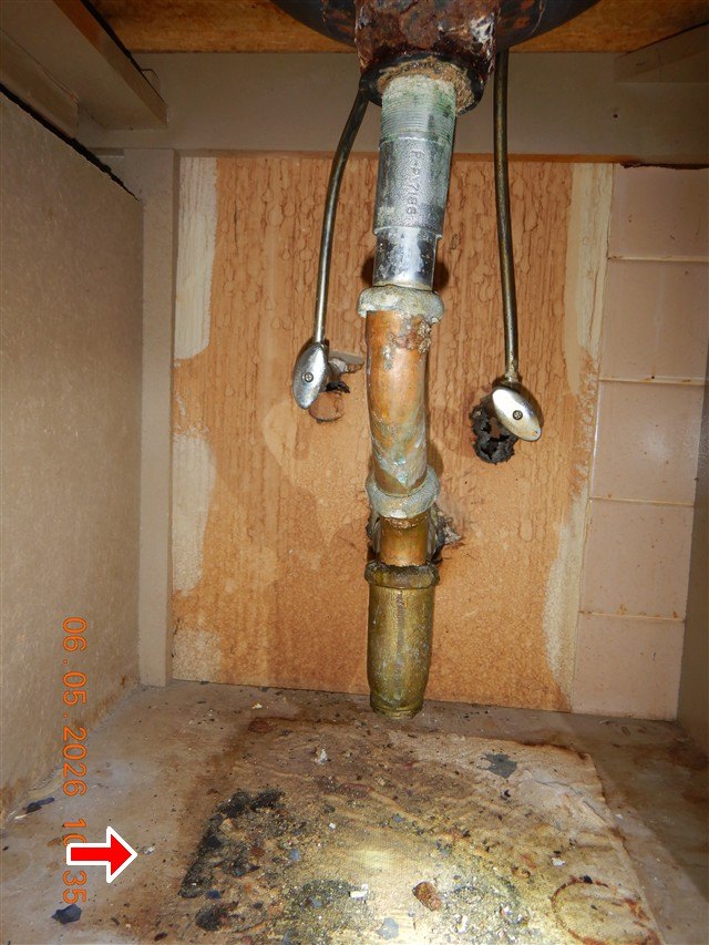 Corroded drain pipe and shut-off valves under a sink, with water staining and mold on the cabinet floor marked by a red arrow.