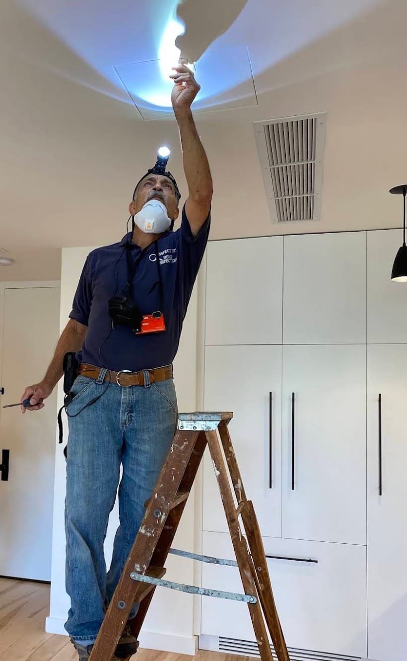 Lou Rubino on a stepladder wearing a headlamp and respirator, reaching up to open a ceiling access hatch during an inspection