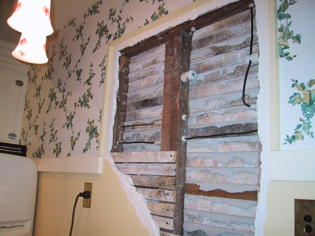 A wall opened to the lath and studs, showing ceramic knobs carrying knob and tube conductors vertically up the stud bay.