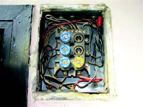An original screw-in fuse panel recessed into a plaster wall, with three blue Edison-base fuses, empty sockets and old cloth-covered conductors.