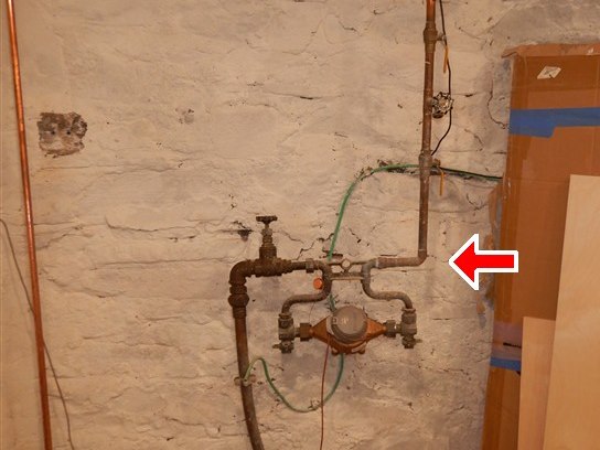 Inspection photograph of a cellar water service entry - shut-off valve, meter and copper pipework against a whitewashed stone foundation wall