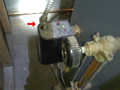 Armored BX cable entering a black control box beside a boiler water feed, with a red arrow at the loose connector.