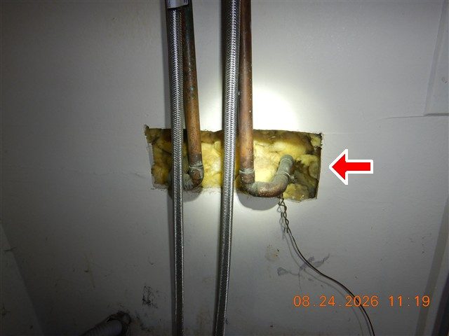 Copper and braided water lines coming through a cut-out in a laundry closet wall with insulation exposed, marked with a red arrow.