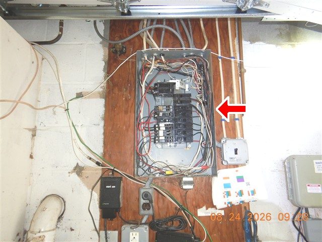 Open main breaker panel on a plywood board in a garage with many wires routed to it, marked with a red arrow.