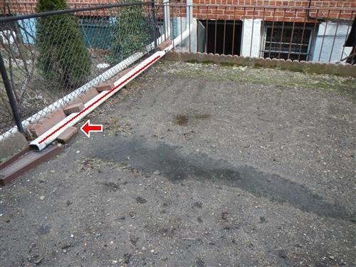 A rear paved yard with a downspout extension running along the fence line, marked with an arrow.