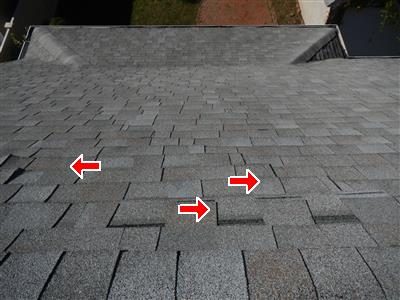 Grey asphalt shingle roof seen from above with three red arrows marking shingles out of line below the peak.