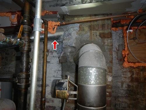 Galvanized flue pipe entering a brick chimney wall beside a stained plaster patch, marked with a red arrow.