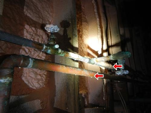 Bare copper heat and water pipes with a valve running along a spray-foamed crawl space wall, two red arrows.