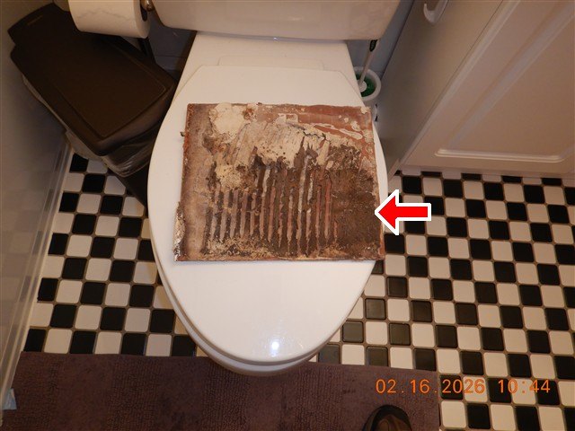 Heavily rusted metal grille with plaster debris resting on a toilet lid, red arrow at corrosion