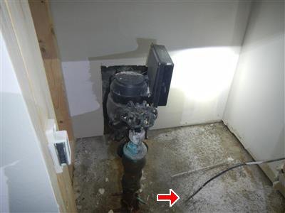 A water service pipe and meter where they enter a house, with staining on the concrete floor around the entry point.