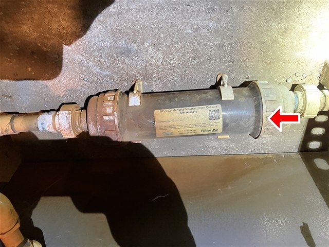 Clear inline condensate neutralizer cartridge installed on piping, red arrow