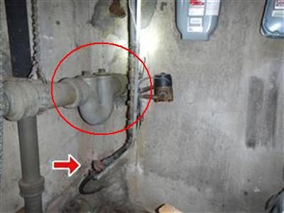 Grey cast iron whole house trap on a basement wall with a red circle around the trap and a red arrow at the storm trap.