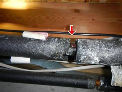 Grey foam-insulated copper pipe under floor joists with a red arrow pointing down at a corroded joint.