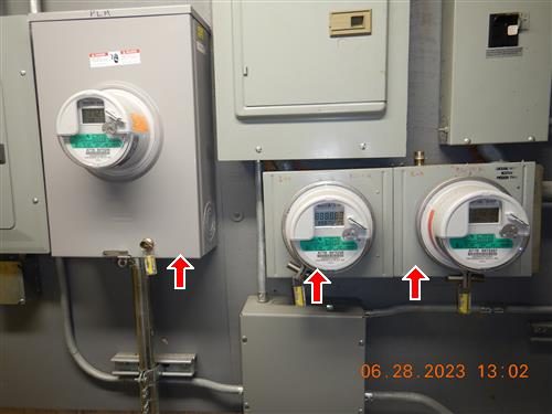 Three electric meters mounted on grey panels on a wall, each marked with a red arrow.