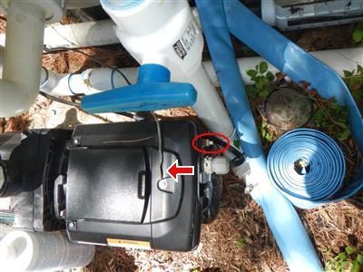 Pool filter pump on the ground with a red arrow and red circle at its base and a blue hose coiled beside it.