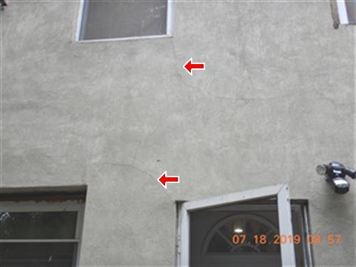 A stucco-parged rear wall with two cracks marked by red arrows beside window openings.