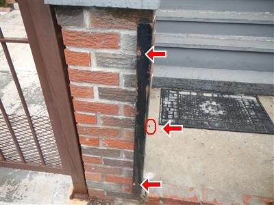 Rusted black steel post set into a brick pier, with three red arrows and a circled hole.
