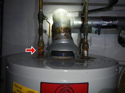 Top of a gas water heater with draft hood and copper piping, red arrow at a corroded fitting.