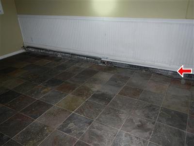 White baseboard heating unit along a wall with staining at its lower edge, red arrow at one end.