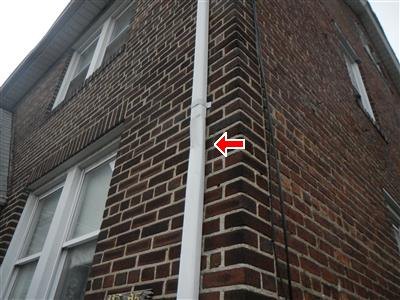 Looking up a brick wall corner at a white downspout, red arrow at a joint in the pipe.
