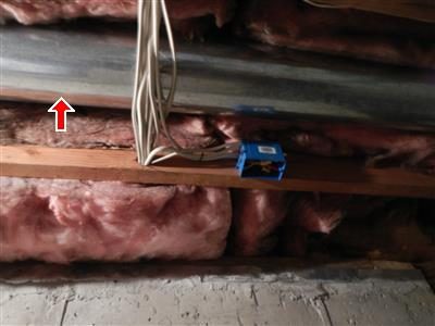 Bare round metal duct pipe running between basement floor joists above pink insulation, marked with a red arrow.