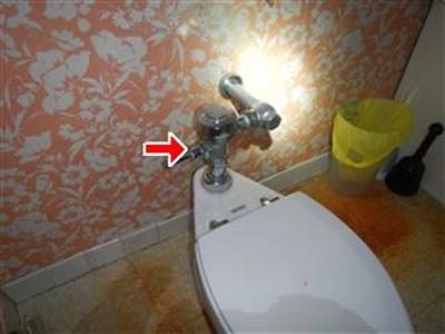Chrome flushometer valve above a toilet tank with a red arrow, stained floor tiles visible below.