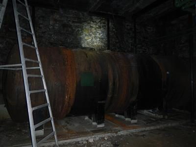Large rusted steel oil tank in a dim basement containment area with a ladder leaning against it.