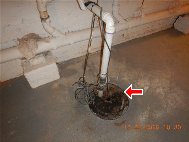 Sump pump pit in a grey basement floor with a white PVC discharge pipe and cords, a red arrow pointing at the pit.