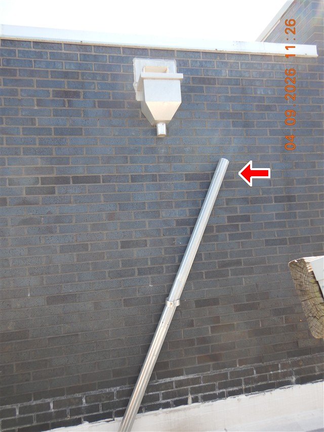 A metal leader pipe leaning disconnected against a dark brick wall beneath its hopper.