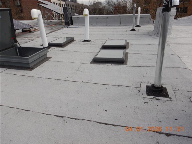 Flat white membrane roof with skylights, a roof hatch, and capped vent pipes, orange date stamp.