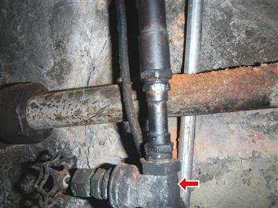 Older steel piping with a valve and fittings against a stone wall, red arrow at one fitting.