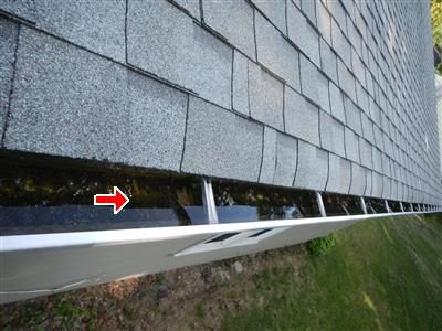 Edge of an asphalt shingle roof with a gutter full of dark debris and water, red arrow at the gutter.