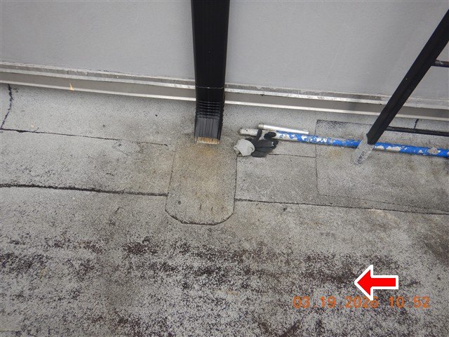 Black downspout ending on a granular flat roof surface with a patched strip, red arrow, date stamp.