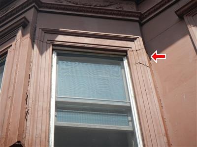 Exterior of a brownstone window with ornate surround, red arrow at cracked material near the frame.