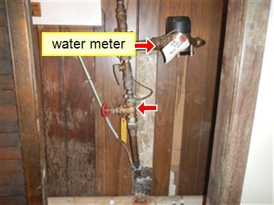 Water supply riser in a basement with a label reading water meter and a red arrow at the red-handled shut-off valve.