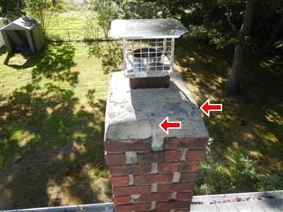 Top of a brick chimney with a rain cap and two red arrows pointing to cracks in the cement cap.