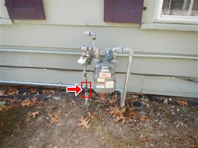 Gas meter against pale siding with a red arrow pointing to the shut-off valve on the riser.