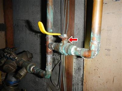 Copper pipes with heavy green corrosion at fittings, yellow valve handle, red arrow at a joint