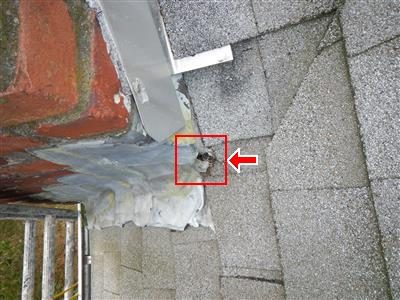 Brick chimney base meeting grey shingles with heavy sealant, a hole in the flashing boxed in red with an arrow.