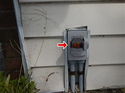 Exterior outlet box on white siding with a red arrow pointing to the receptacle, two grey conduits running down below it.