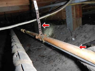 Bare copper water pipe in a crawl space held by a perforated steel strap, red arrows at the strap and the pipe.