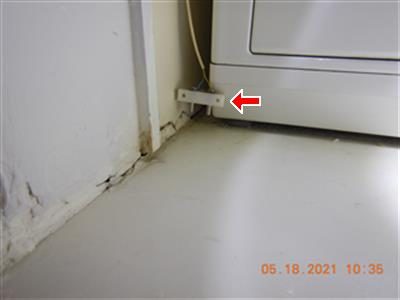 Small white water-alarm sensor beside a clothes washer, held above the floor, with a red arrow pointing at it.