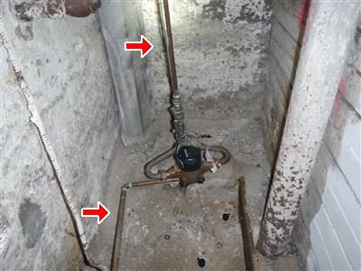 Water meter on a narrow pipe in a basement corner with red arrows on the pipe above and below it.