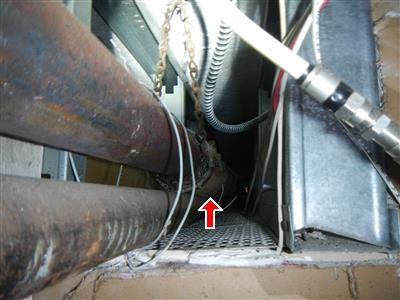 Basement ceiling enclosure with a rusted steam pipe and flexible metal conduit, a red arrow pointing to the insulation at the pipe joint.