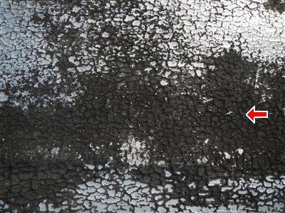 Flat roof surface with cracked, alligatored black coating over silver paint, red arrow