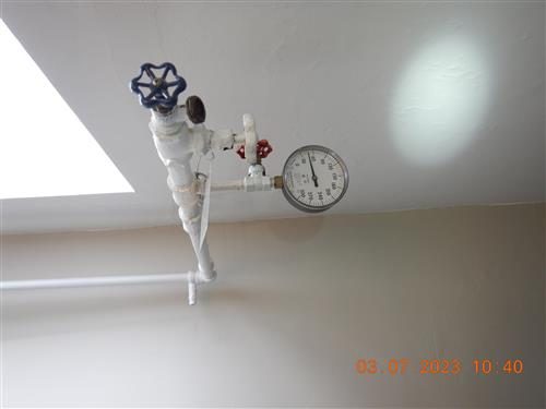 Pressure gauge fitted to a white supply pipe and valve on a top-floor wall.
