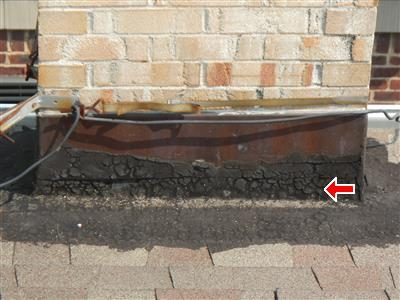 Base of a tan brick chimney where it meets a shingle roof, with a rusted metal strip and cracked black cement, red arrow at the right.