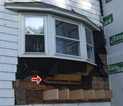 Bay window with siding removed, exposing black felt, damaged sheathing and new lumber, red arrow on framing