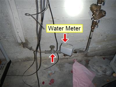 Water meter and shut-off valve on a basement supply pipe, marked with a yellow label and arrows